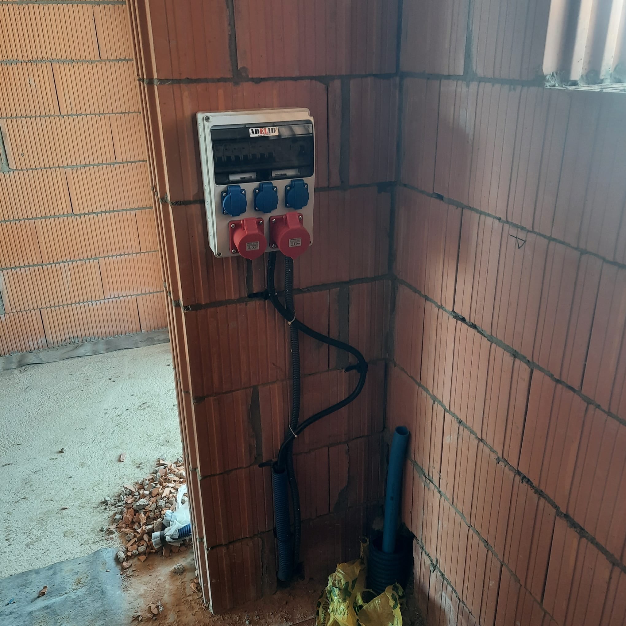 Rozdzielnia elektryczna z gniazdami siłowymi na ścianie z czerwonej cegły dziurawki, kable w peszlach. Instalacja w trakcie montażu w surowym stanie.