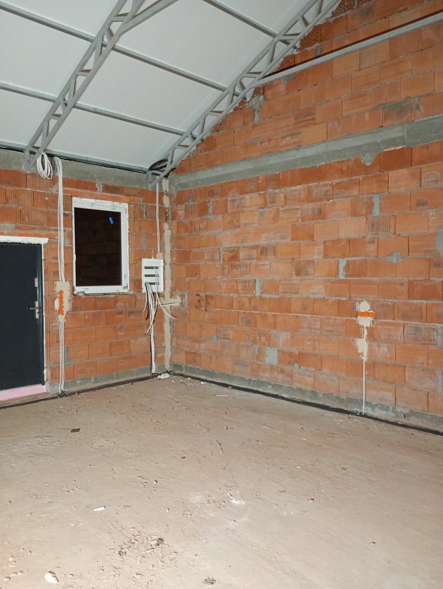 Wnętrze surowego pomieszczenia z instalacją elektryczną: kable prowadzone po ścianach z cegły, skrzynka rozdzielcza pod oknem. Widoczna stalowa konstrukcja dachu.