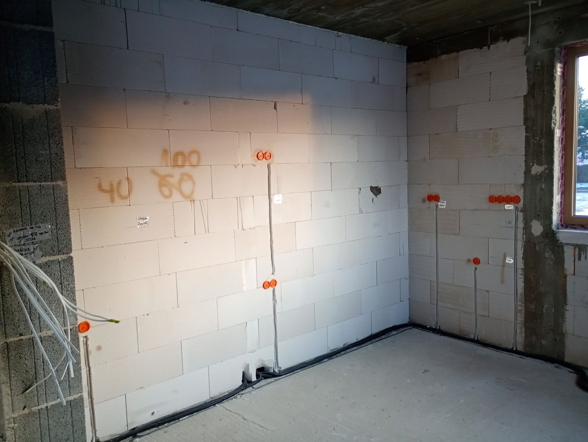 Surowa ściana z betonu komórkowego z instalacją elektryczną w trakcie montażu; widoczne kable, puszki i oznaczenia. W tle okno i fragment sufitu.