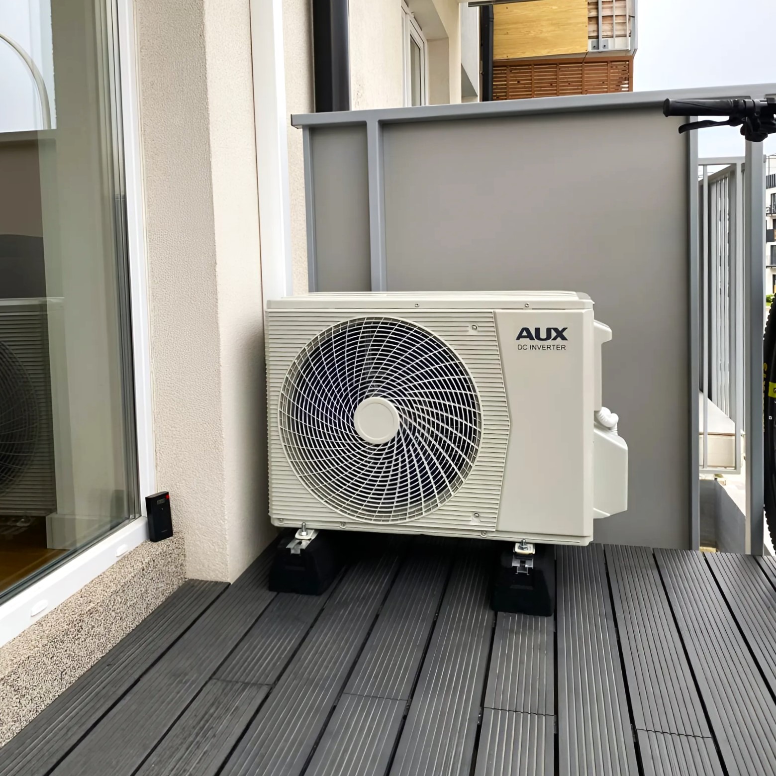 Biała jednostka zewnętrzna klimatyzacji AUX DC Inverter na balkonie z szarą podłogą z desek kompozytowych i szarą barierką. Widok z boku, lekko z dołu.