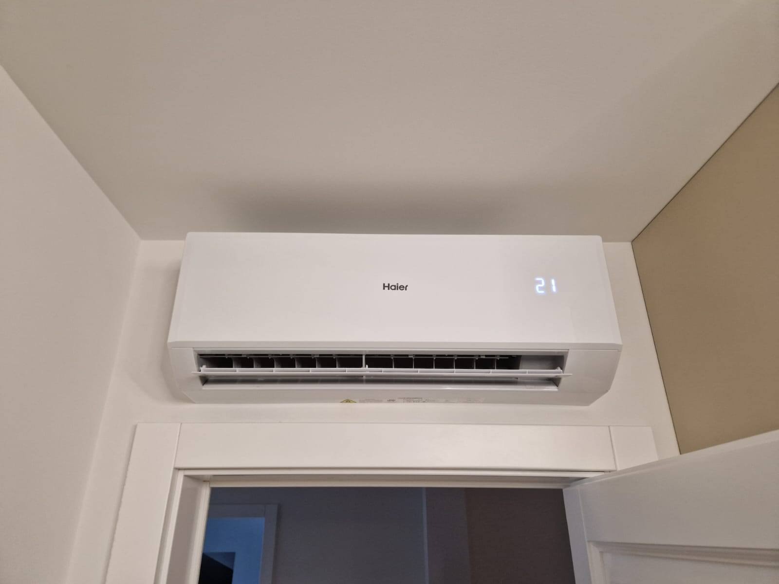Biała klimatyzacja Haier z wyświetlaczem 21 stopni, zamontowana nad drzwiami w jasnym wnętrzu.