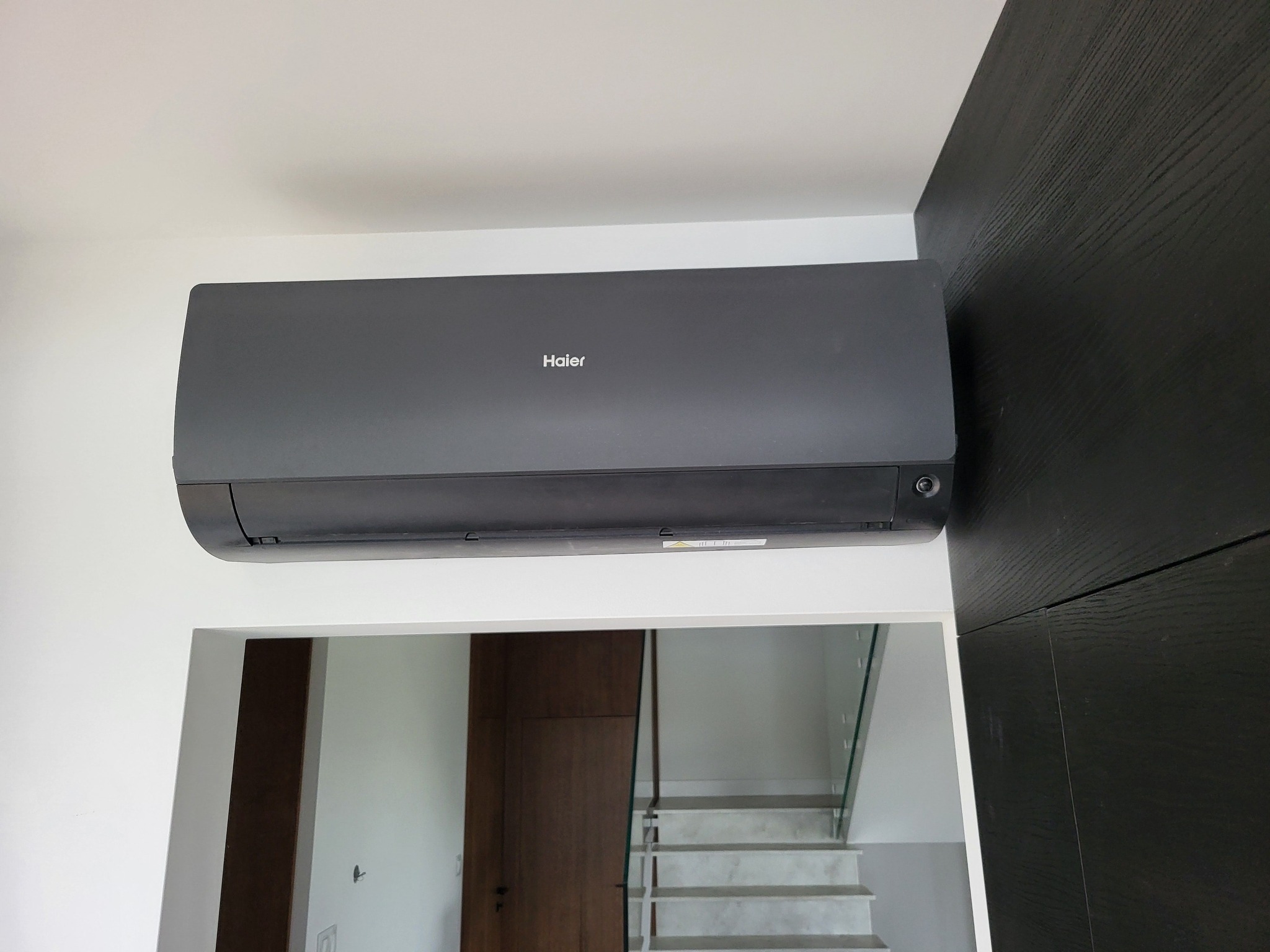 Czarny klimatyzator Haier zamontowany na białej ścianie, z widocznym fragmentem czarnej ściany z drewnianą fakturą. W tle schody z przeszkloną balustradą.
