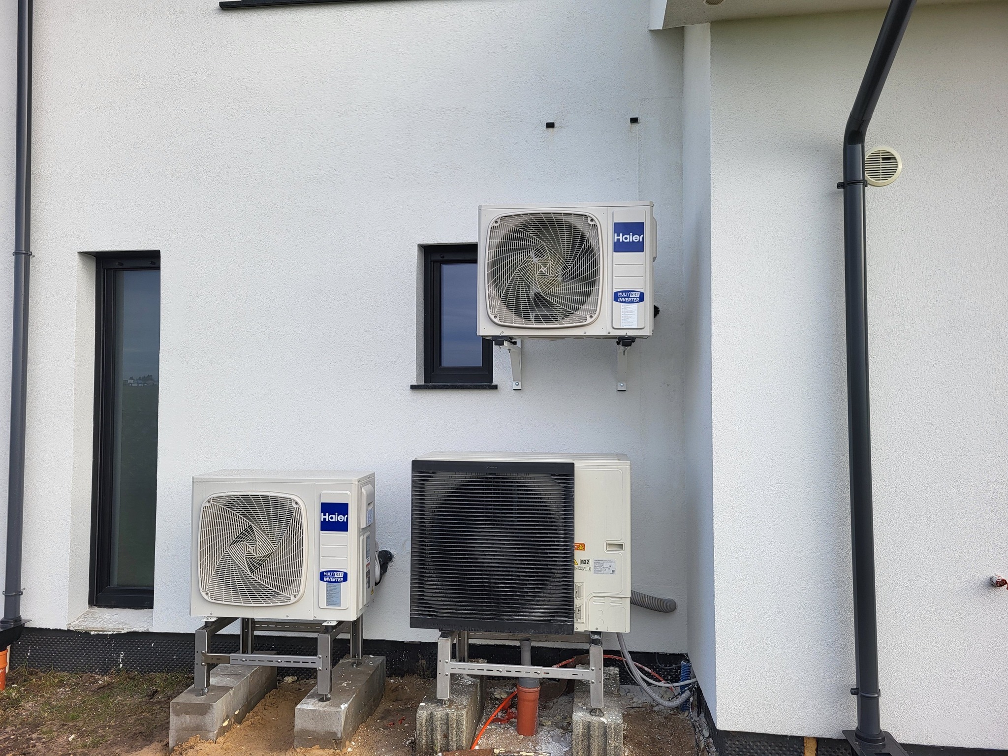 Biała ściana budynku z trzema jednostkami zewnętrznymi klimatyzacji Haier, każda zamontowana na metalowych wspornikach. Widoczne okno i rura spustowa.
