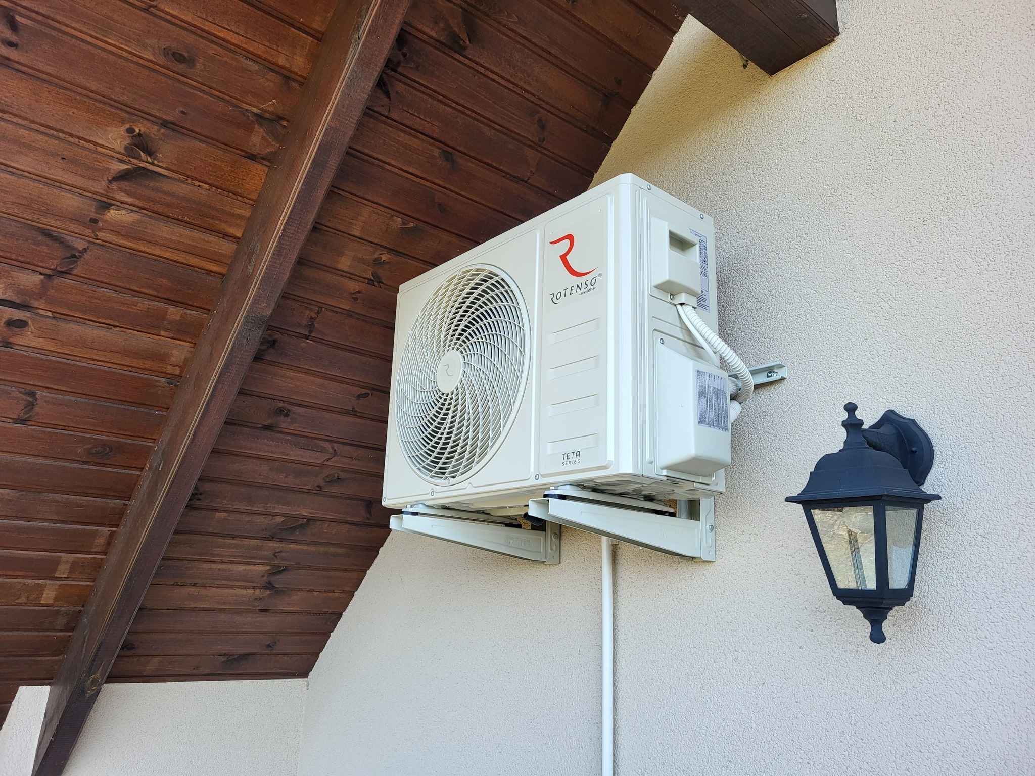 Zamontowany klimatyzator Rotenso Teta Series na elewacji domu z drewnianym podbitką i ozdobną lampą ścienną. Widoczne uchwyty montażowe i przewody.
