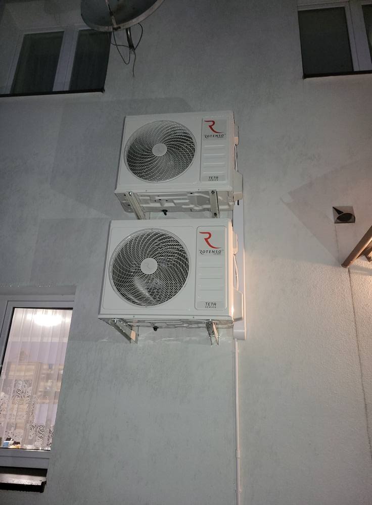 Dwa białe klimatyzatory Rotenso Teta Series zamontowane jeden nad drugim na szarej ścianie budynku, widoczne okno i antena satelitarna.