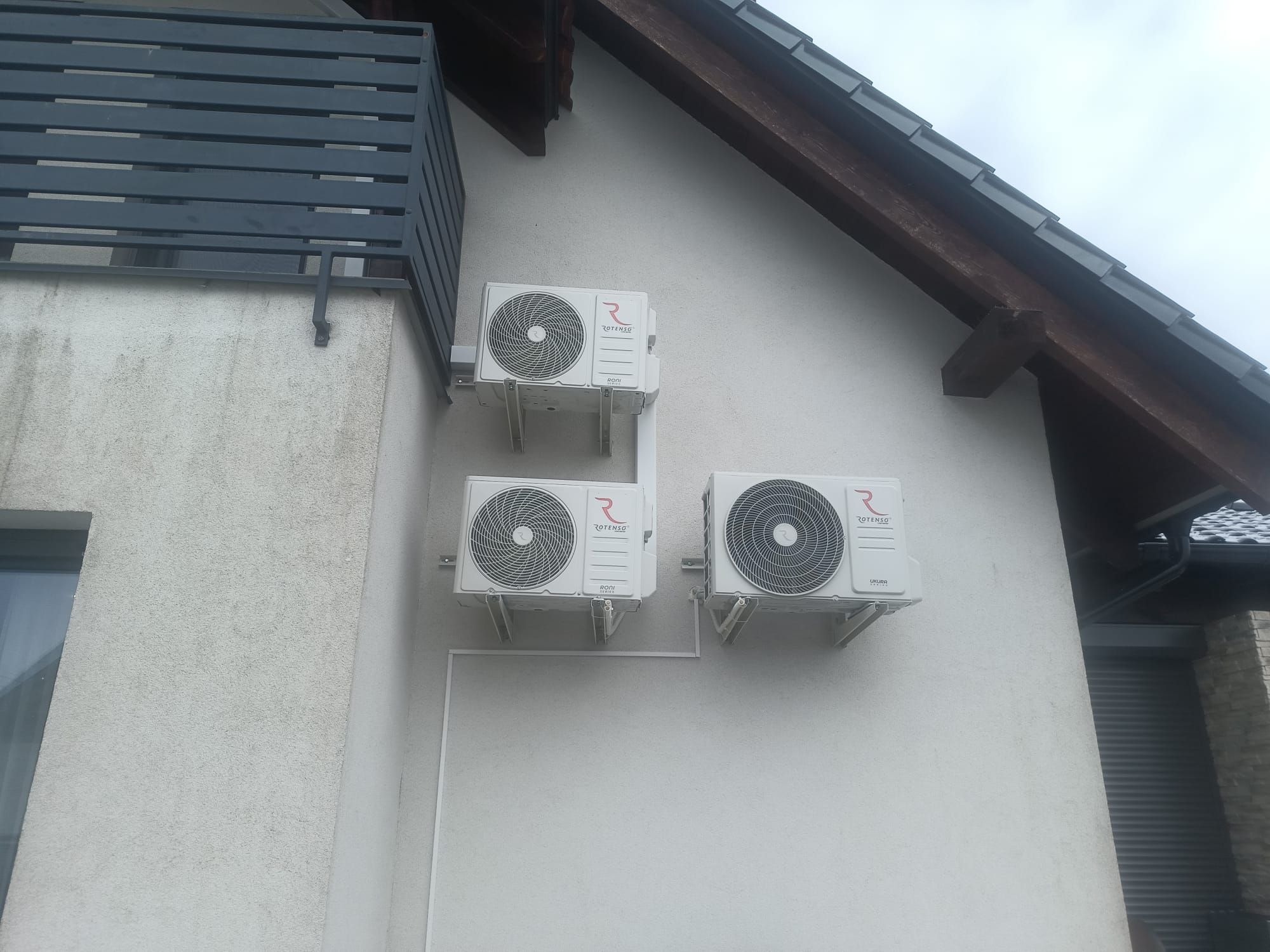 Trzy białe klimatyzatory Rotenso zamontowane na elewacji domu, połączone przewodami. Widoczny fragment dachu z ciemną dachówką i balkon z szarą balustradą.