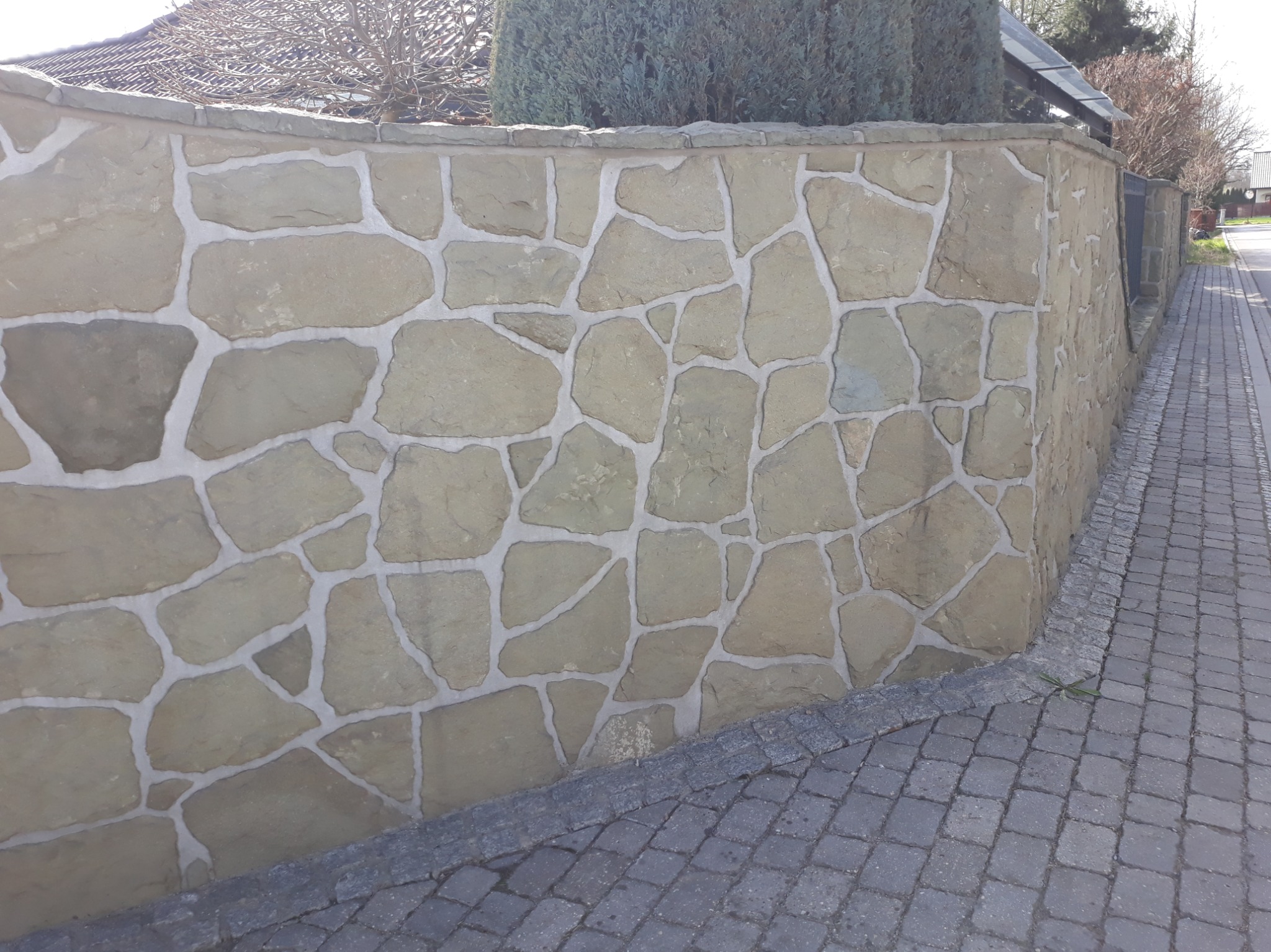 Kamienny mur ogrodzeniowy z nieregularnych piaskowcowych bloków, spoinowany jasną zaprawą, przylegający do kostki brukowej chodnika. W tle widoczne krzewy i fragment domu.