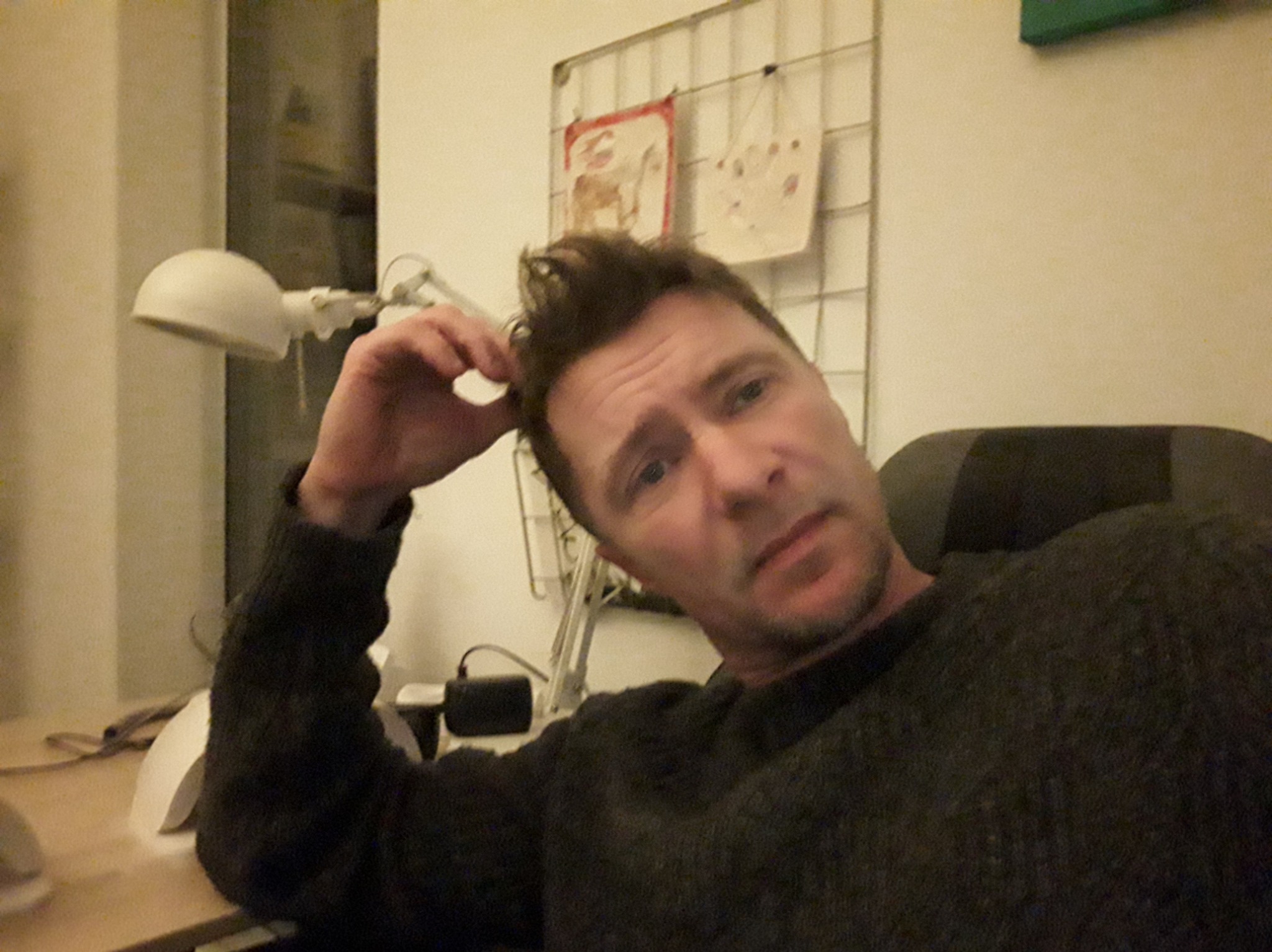 Mężczyzna w ciemnym swetrze siedzi przy biurku, opiera głowę na dłoni, w tle lampka biurkowa i siatka z dziecięcymi rysunkami.