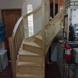 Jasne, drewniane schody kręte z balustradą wewnątrz domu, widoczne na tle ściany z luksferami i drewnianych drzwi. Elementy wykończeniowe w surowym drewnie.