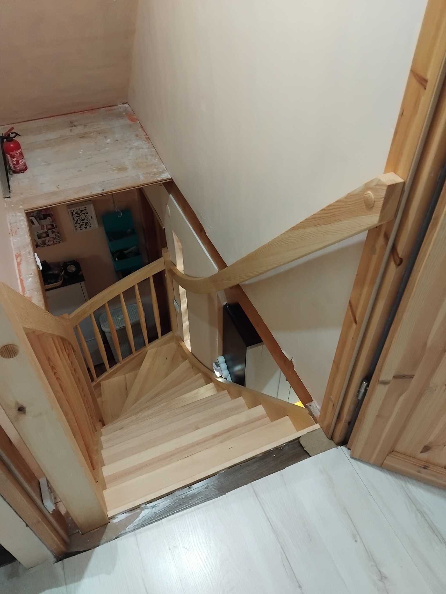 Jasne, drewniane schody z barierką, widziane z góry, prowadzą na niższy poziom z widocznym fragmentem kuchni i gaśnicą. Oświetlenie dzienne.