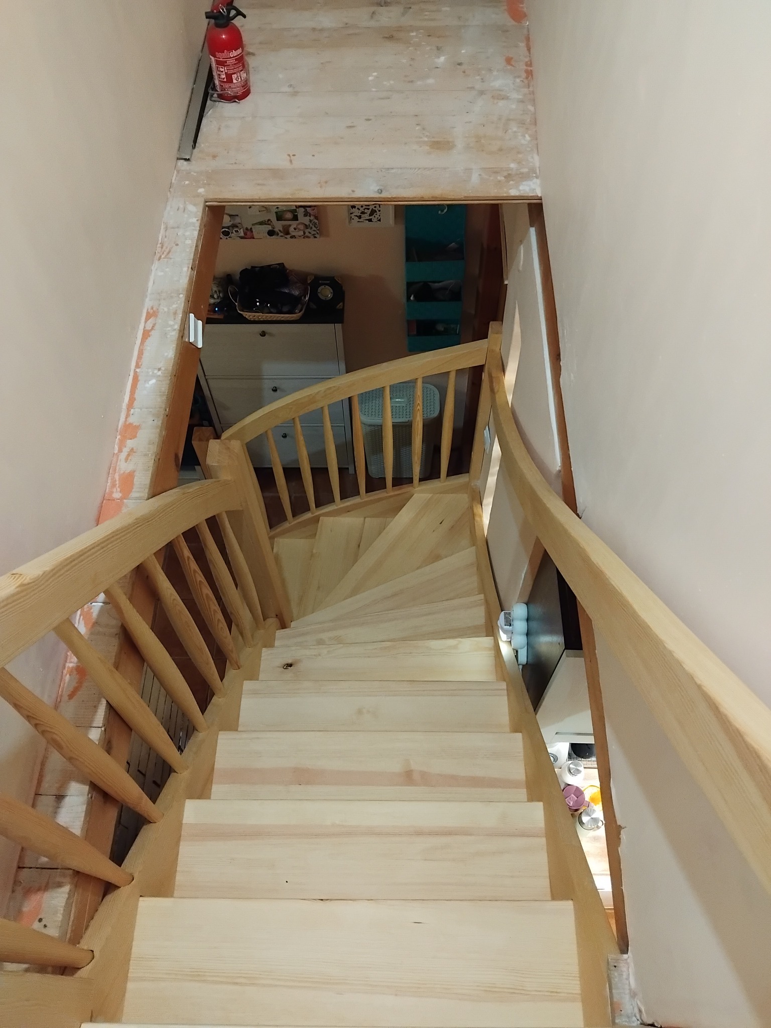 Wysoki kąt: Jasne, spiralne schody z surowego drewna prowadzą w dół, widoczne niedokończone prace wykończeniowe i gaśnica na górze. Widok na wnętrze domu.