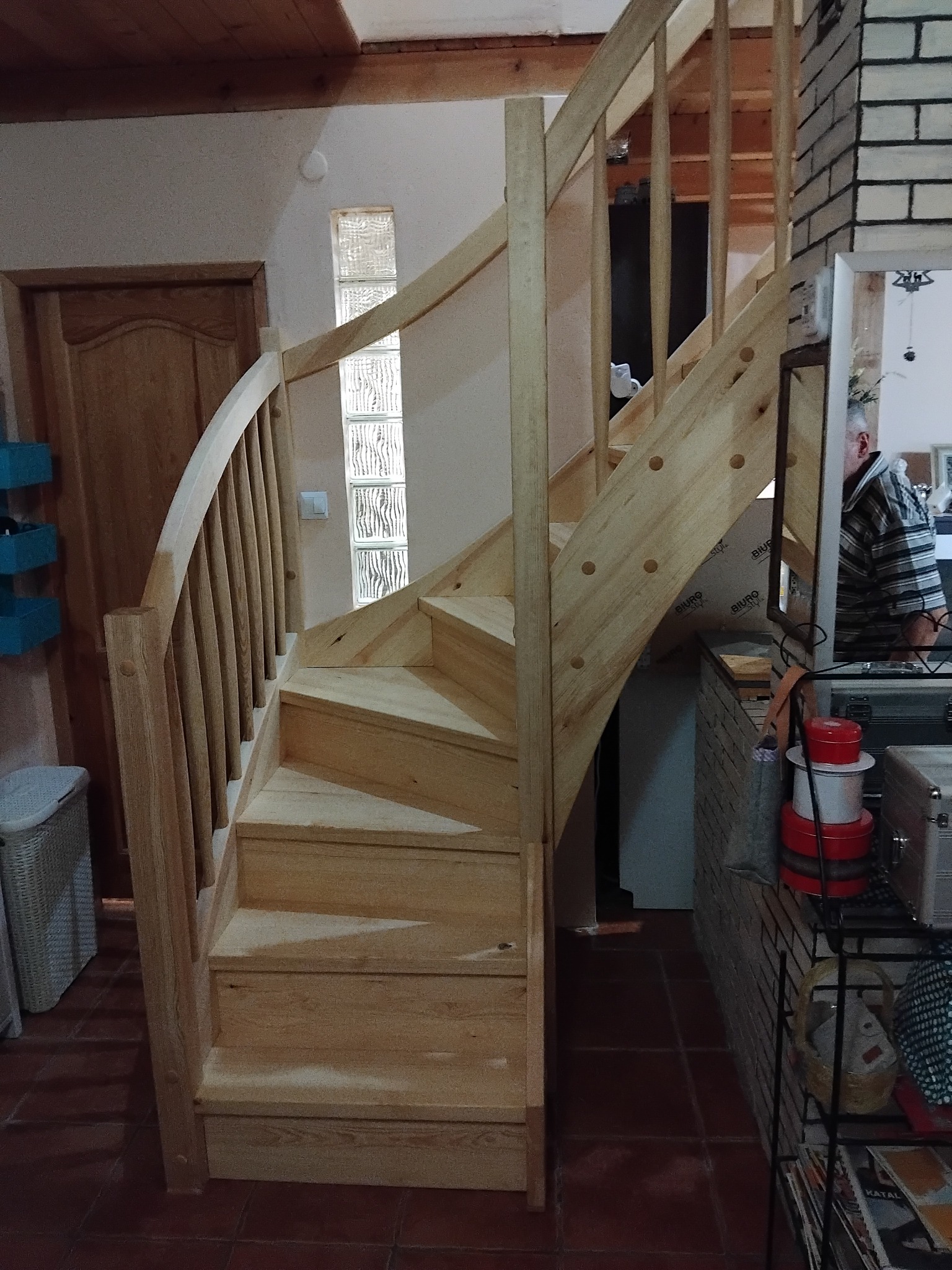 Jasne, drewniane schody kręte z balustradą wewnątrz domu, widoczne na tle ściany z luksferami i drewnianych drzwi. Elementy wykończeniowe w surowym drewnie.