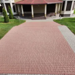 Czerwona kostka brukowa na podjeździe w Koszalinie, ułożona w regularny wzór. Widoczne obrzeża z szarej kostki, trawnik i fragment budynku w tle.
