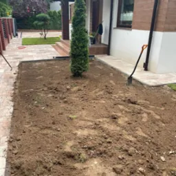 Przygotowany teren pod trawnik z widoczną tujką, obrzeżami i narzędziami ogrodniczymi. Dom z kostką brukową w tle. Grunt świeżo przekopany, gotowy do siewu lub rozłożenia trawy.