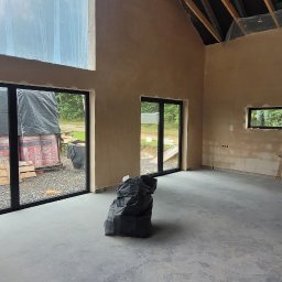 Mach-Tynk - Surowy stan wykończenia wnętrza w Żywcu: ściany po wstępnym nałożeniu tynku, widoczne ślady stóp na szarym podłożu i czarny worek gruzu na środku pomieszczenia.