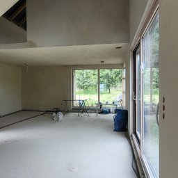 Mach-Tynk - Wnętrze w trakcie prac wykończeniowych: ściany pokryte świeżym tynkiem maszynowym, widoczne okno z widokiem na zieleń, niebieski worek i narzędzia na podłodze.