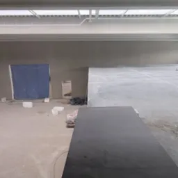 Wnętrze w trakcie prac wykończeniowych. Widoczna ściana po nałożeniu gładzi, sufit z instalacjami oraz podłoże przygotowane do dalszych prac. W tle zasłonięte wejście.