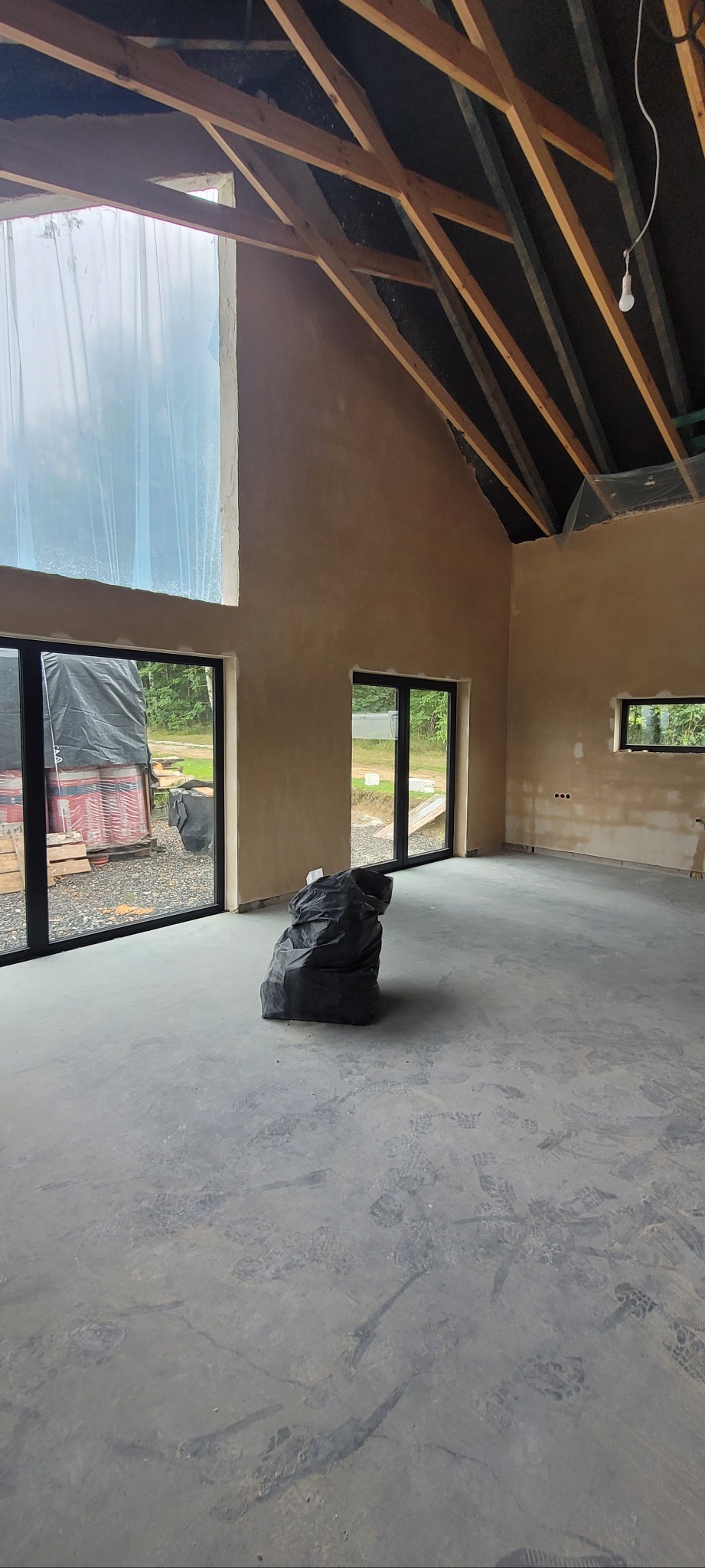 Surowy stan wykończenia wnętrza w Żywcu: ściany po wstępnym nałożeniu tynku, widoczne ślady stóp na szarym podłożu i czarny worek gruzu na środku pomieszczenia.