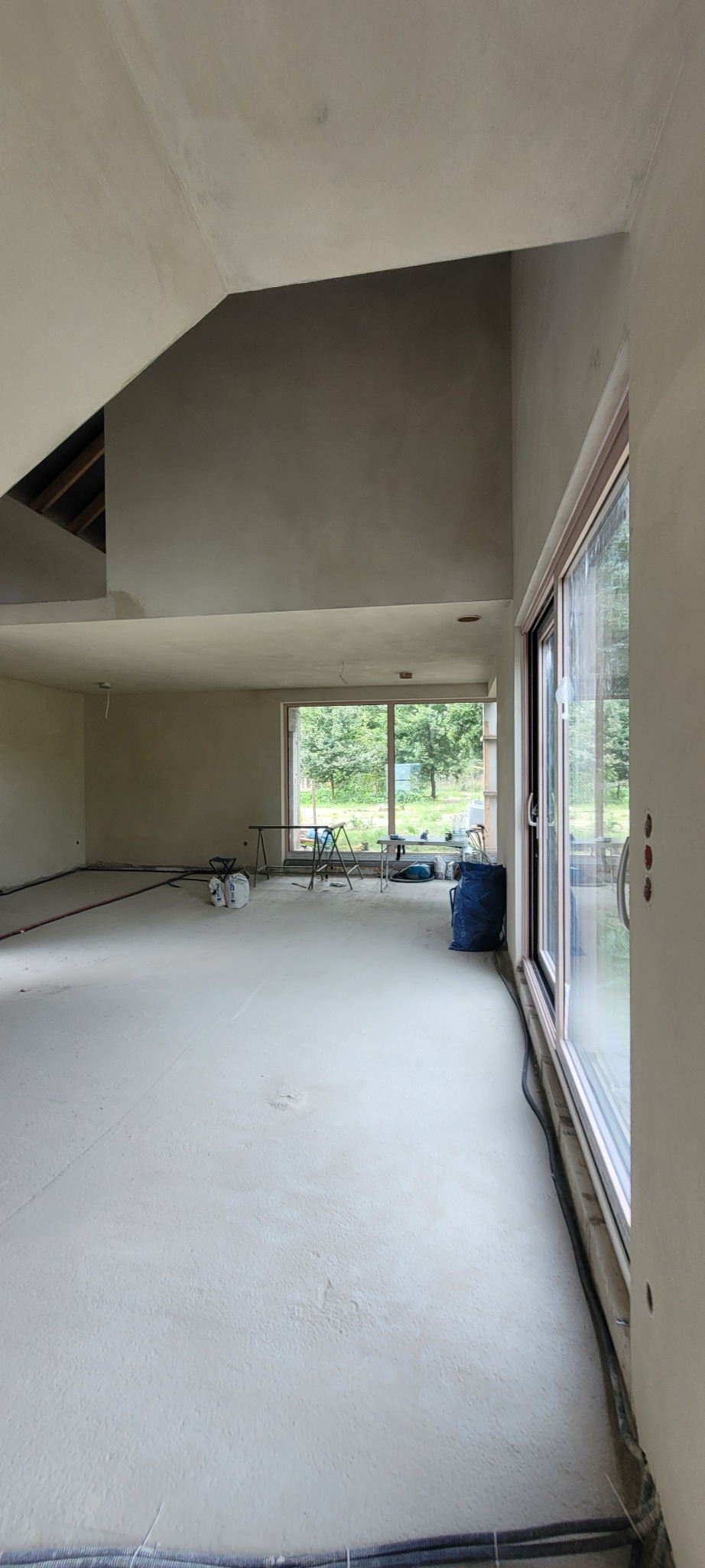 Wnętrze w trakcie prac wykończeniowych: ściany pokryte świeżym tynkiem maszynowym, widoczne okno z widokiem na zieleń, niebieski worek i narzędzia na podłodze.