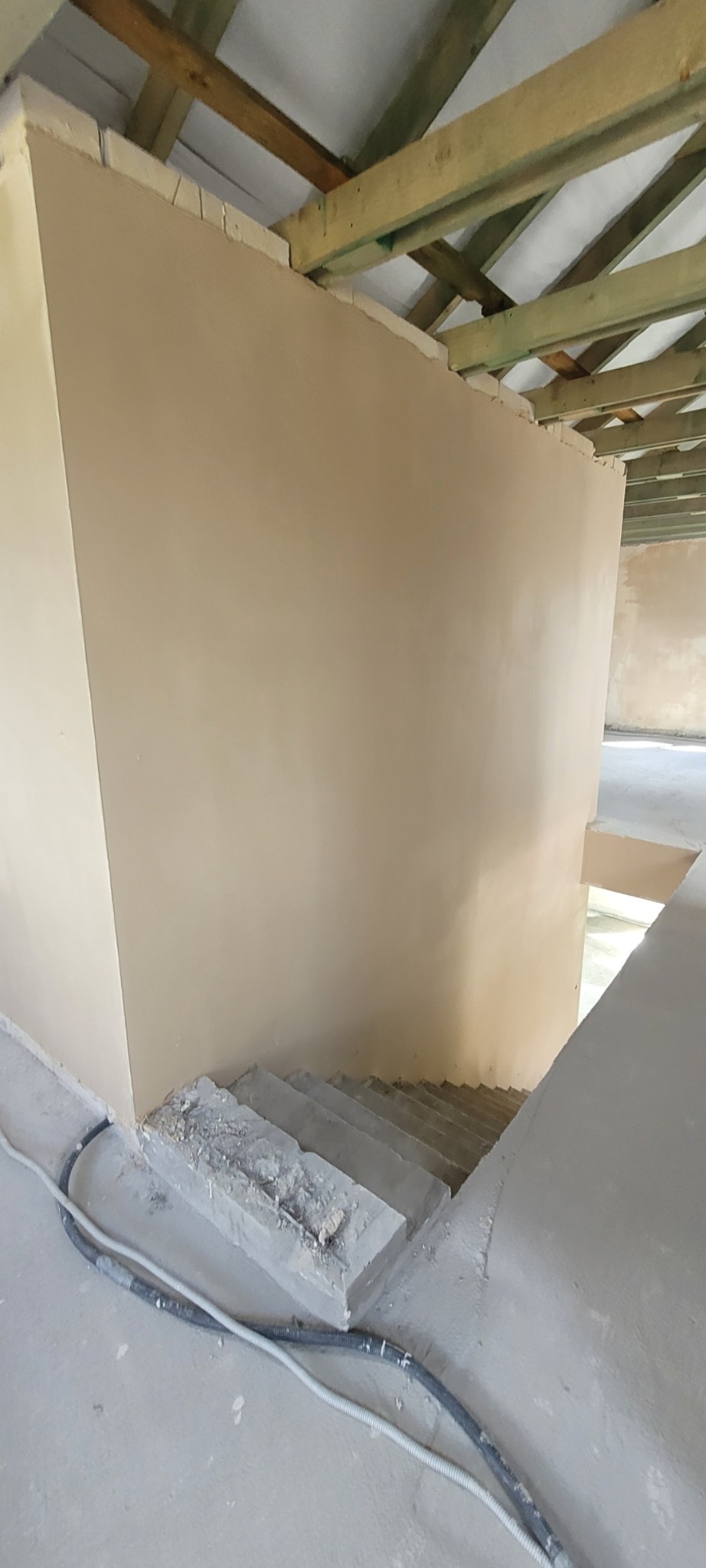 Świeżo położona, beżowa gładź na ścianie w surowym wnętrzu. Widoczne drewniane elementy konstrukcji dachu i betonowe schody. Na podłodze leżą kable w peszlu.