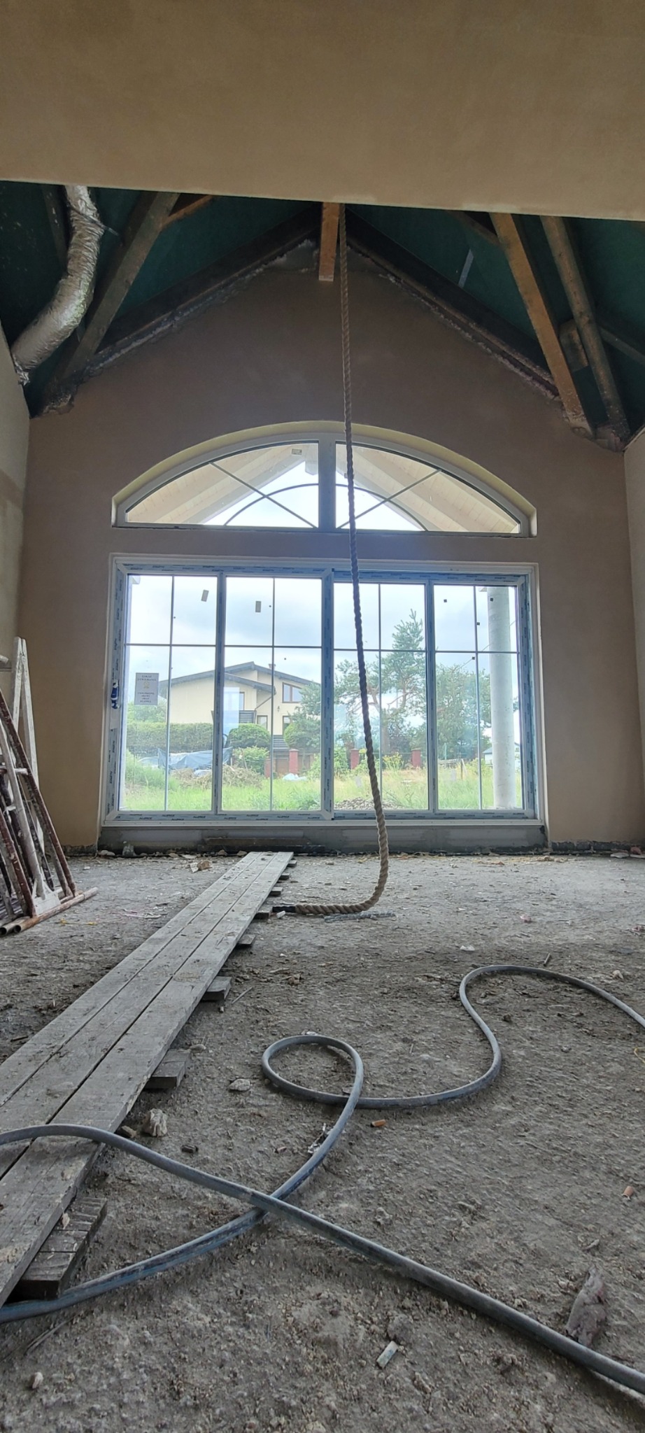 Wnętrze z dużym oknem po nałożeniu tynku. Widoczne drewniane elementy konstrukcji dachu, szare podłoże z przewodami. Uwagę przykuwa lina zwisająca z sufitu.