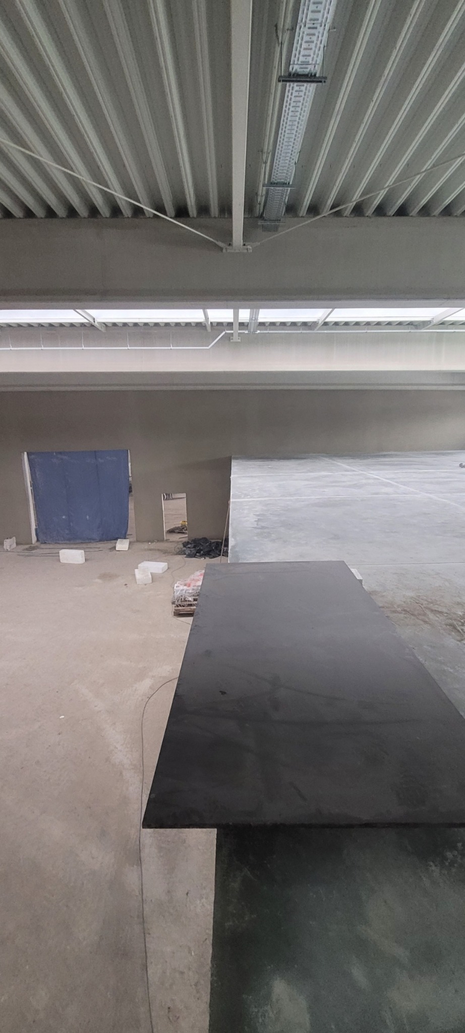 Wnętrze w trakcie prac wykończeniowych. Widoczna ściana po nałożeniu gładzi, sufit z instalacjami oraz podłoże przygotowane do dalszych prac. W tle zasłonięte wejście.