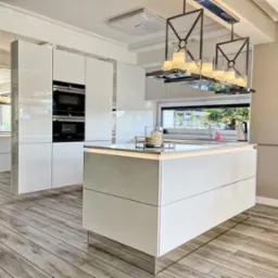 Nowoczesna, jasna kuchnia z wyspą i oświetleniem LED, fronty szafek na wysoki połysk, drewnopodobna podłoga i designerskie lampy wiszące.