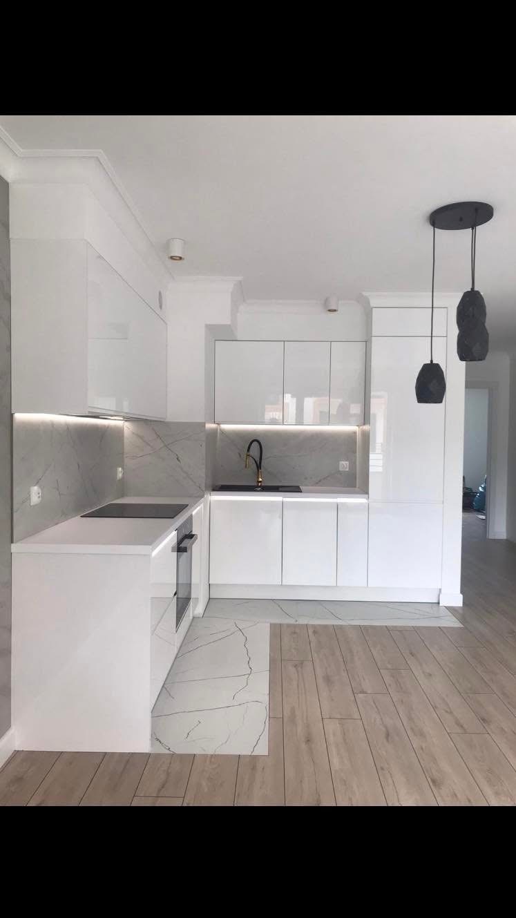 Biała kuchnia na wymiar z marmurowym blatem i podświetleniem LED. Czarne elementy armatury i lampy nadają nowoczesny akcent. Jasna podłoga imitująca drewno.