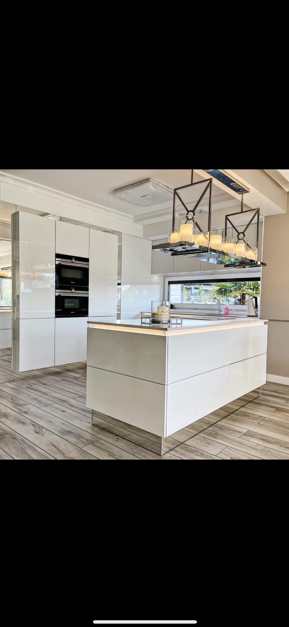 Biała, nowoczesna kuchnia z wyspą i oświetleniem LED, wykończona elementami lustrzanymi. Widoczne zabudowane sprzęty i drewnopodobna podłoga.