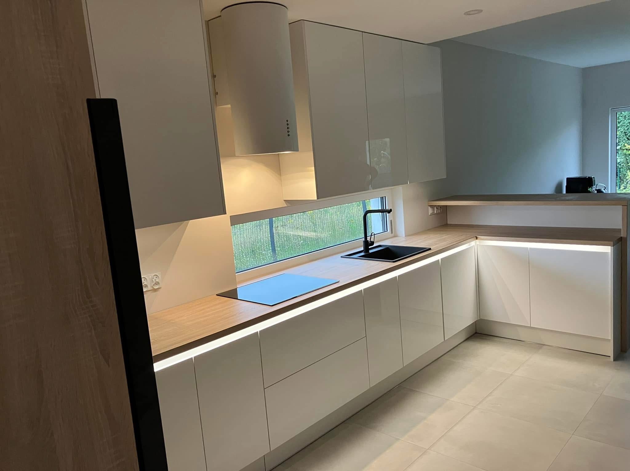 Nowoczesna kuchnia z białymi, lakierowanymi frontami, drewnianym blatem i podświetleniem LED. Widok na okno z czarnym zlewem i płytą grzewczą.