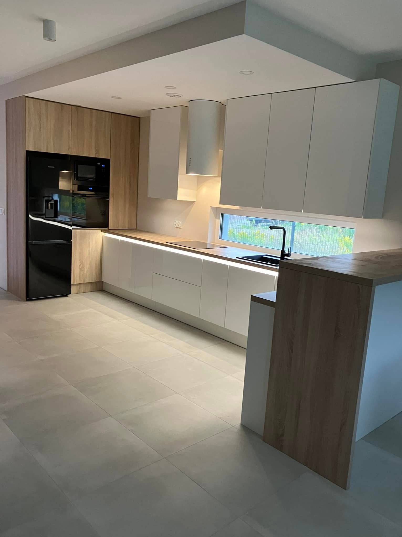 Nowoczesna kuchnia z białymi szafkami i drewnianymi akcentami, oświetlona listwami LED pod szafkami wiszącymi. Widok na ogród przez okno nad zlewem.