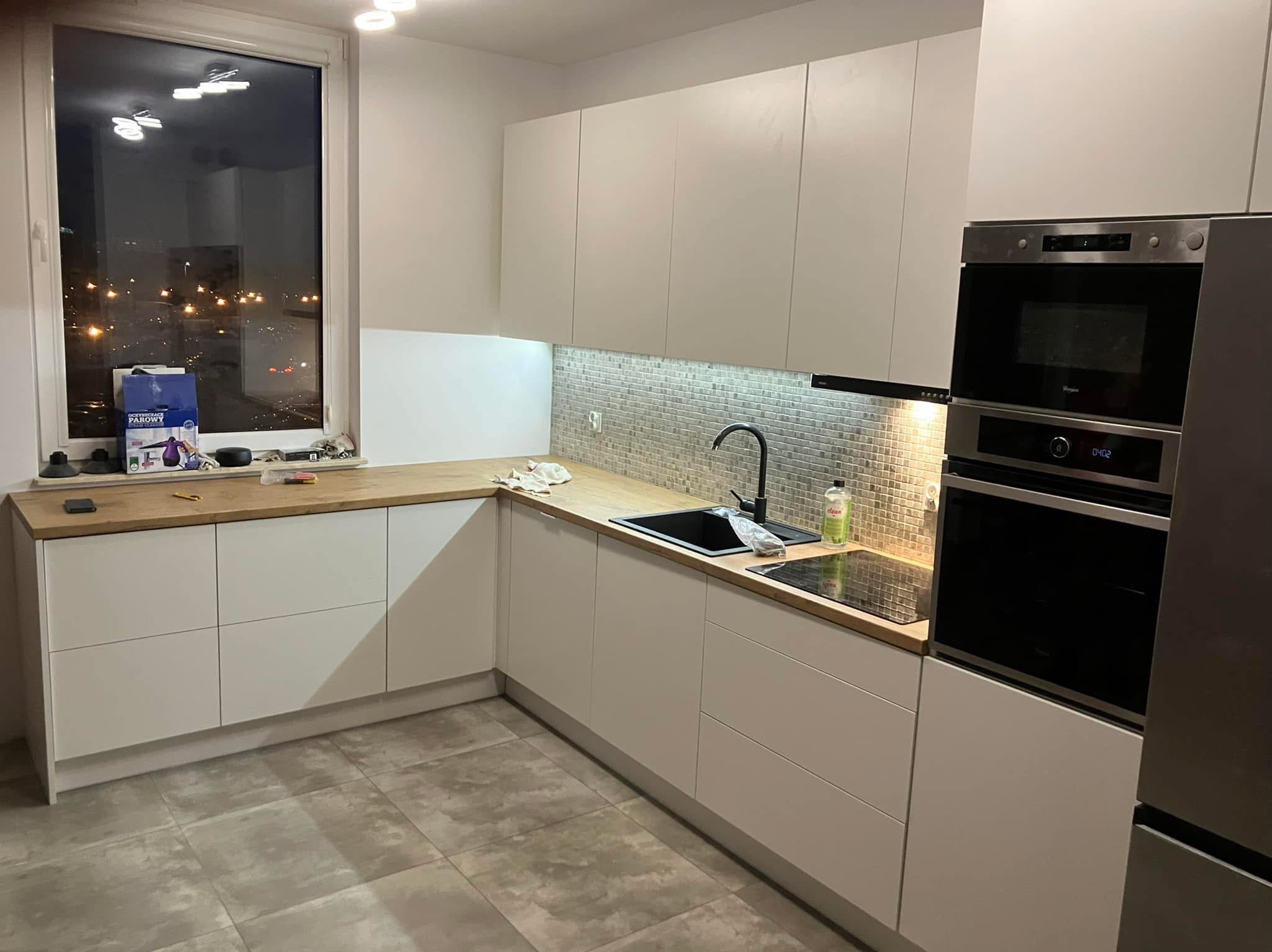 Nowoczesna kuchnia z białymi frontami, drewnianym blatem i mozaiką nad blatem. Widok na okno z widokiem na miasto nocą. Nowe sprzęty AGD w zabudowie.