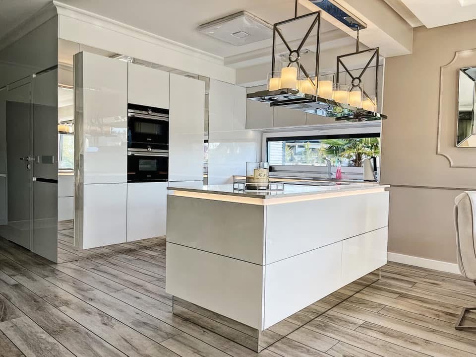 Nowoczesna, jasna kuchnia z wyspą i oświetleniem LED, fronty szafek na wysoki połysk, drewnopodobna podłoga i designerskie lampy wiszące.