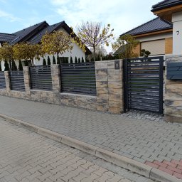 MUROWANE KARILO - Nowoczesne ogrodzenie z paneli i kamienia łupanego, brama wejściowa, skrzynka na listy z numerem domu na tle dwóch budynków mieszkalnych.