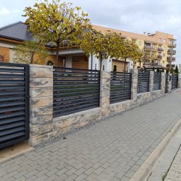 MUROWANE KARILO - Nowoczesne ogrodzenie panelowe z elementami betonowymi i bramą, z przesuwnymi poziomymi panelami, na tle domu jednorodzinnego i bloku mieszkalnego.