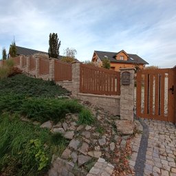 MUROWANE KARILO - Drewniana brama wejściowa z kamiennymi słupkami, prowadząca do domu z drewnianą elewacją. Uwagę przykuwa skrzynka na listy w stylu retro i ozdobna kostka brukowa.