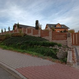 MUROWANE KARILO - Drewniano-kamienne ogrodzenie posesji z bramą wjazdową, na tle drewnianych domów. Kątowe ujęcie z poziomu chodnika z kostki brukowej.