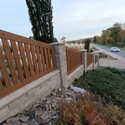 MUROWANE KARILO - Drewniano-betonowe ogrodzenie posesji z elementami dekoracyjnymi z kamienia i roślinności. W tle droga z jadącym samochodem i drzewa.