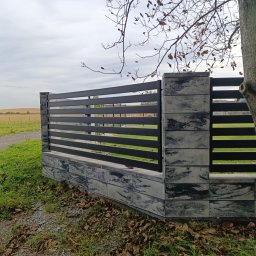 MUROWANE KARILO - Nowoczesne ogrodzenie betonowe z poziomymi, czarnymi przęsłami, widok z boku. Tło: zielone pole i drzewa. Fragment pnia drzewa w prawym dolnym rogu kadru.