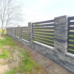 MUROWANE KARILO - Nowoczesne ogrodzenie z szarego betonu architektonicznego z poziomymi, metalowymi przęsłami. Widok na fragment posesji i drzewo w tle.