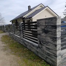 Nowoczesne ogrodzenie z betonowych bloczków w odcieniach szarości i czarnych, poziomych przęseł, na tle domu z ciemną dachówką w pochmurny dzień.