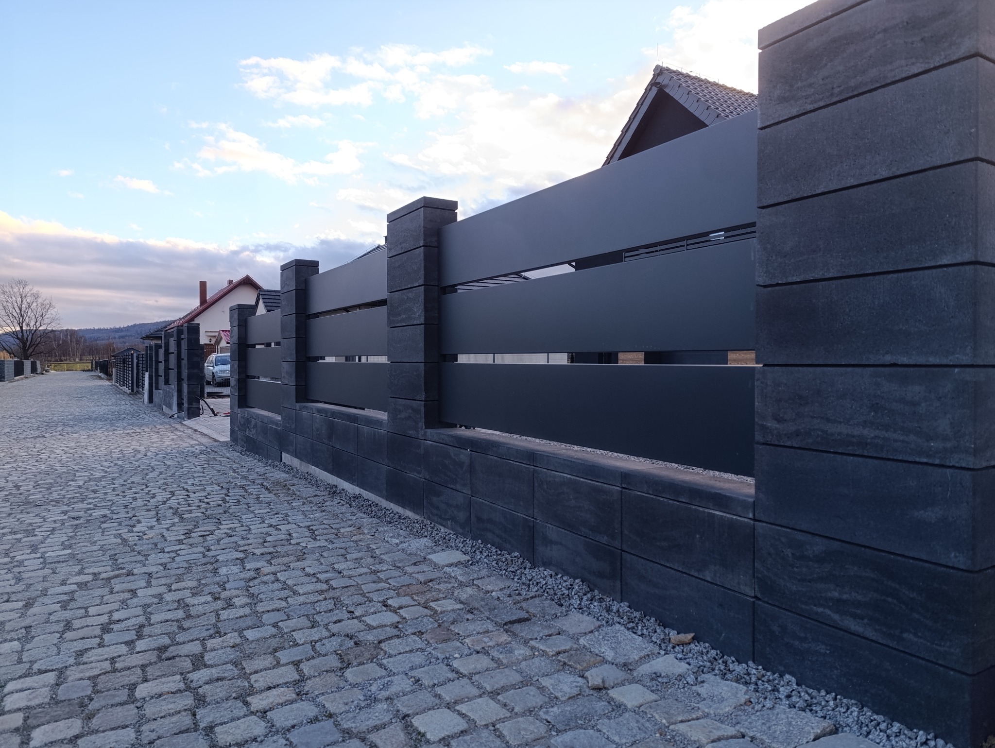 Nowoczesne ogrodzenie panelowe z betonową podmurówką i słupkami, widok z perspektywy ulicy wyłożonej kostką brukową, dom w tle.