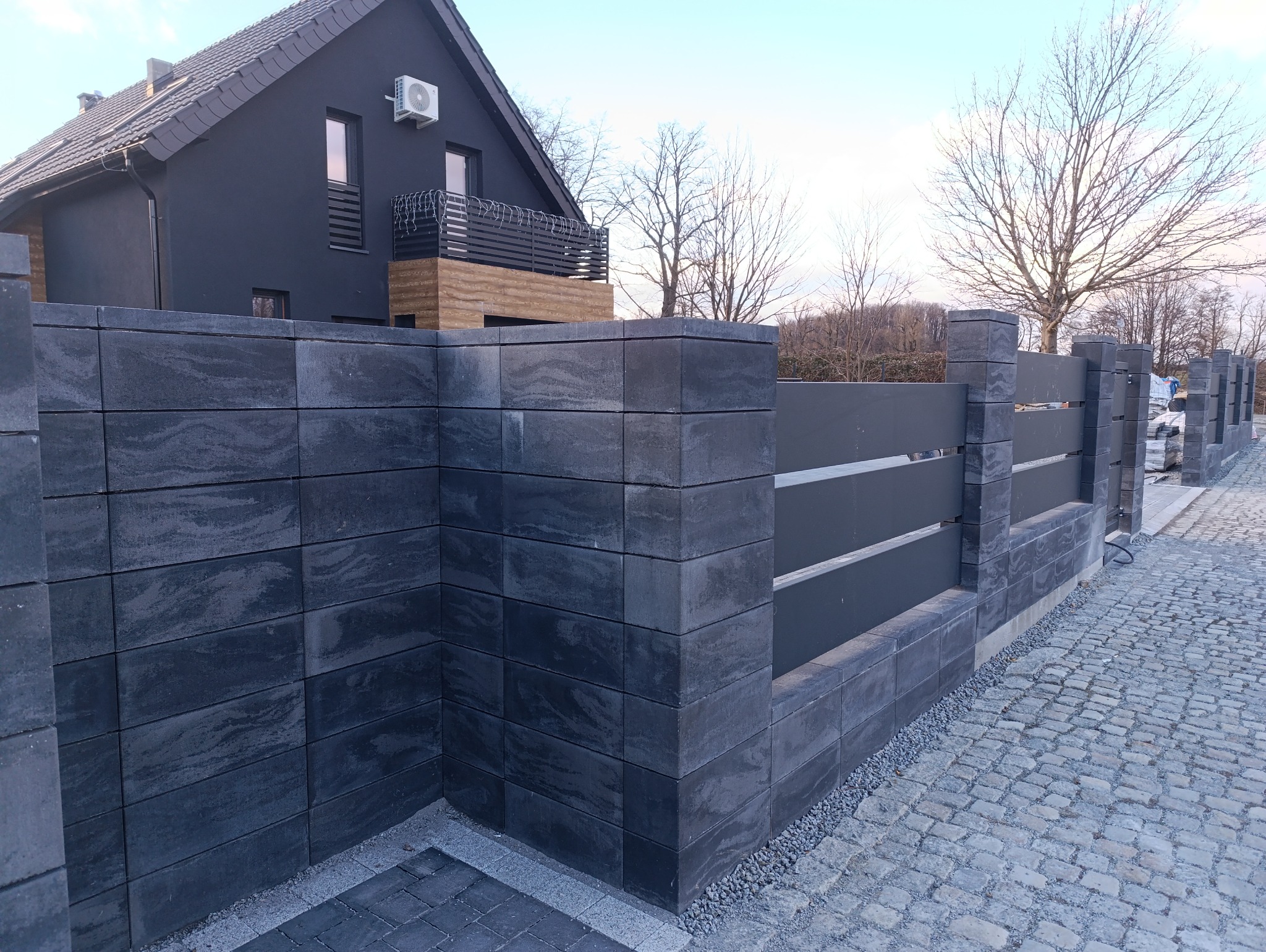 Nowoczesne ogrodzenie z szarych, betonowych bloczków i metalowych paneli, z widocznym fragmentem domu w tle. Estetyczne wykończenie posesji.