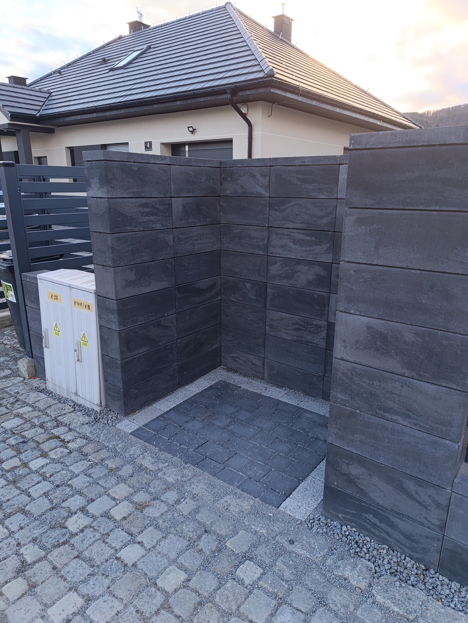 Nowoczesne, grafitowe ogrodzenie modułowe z betonu architektonicznego, częściowo wzniesione, z widocznym fragmentem posesji i kostką brukową.