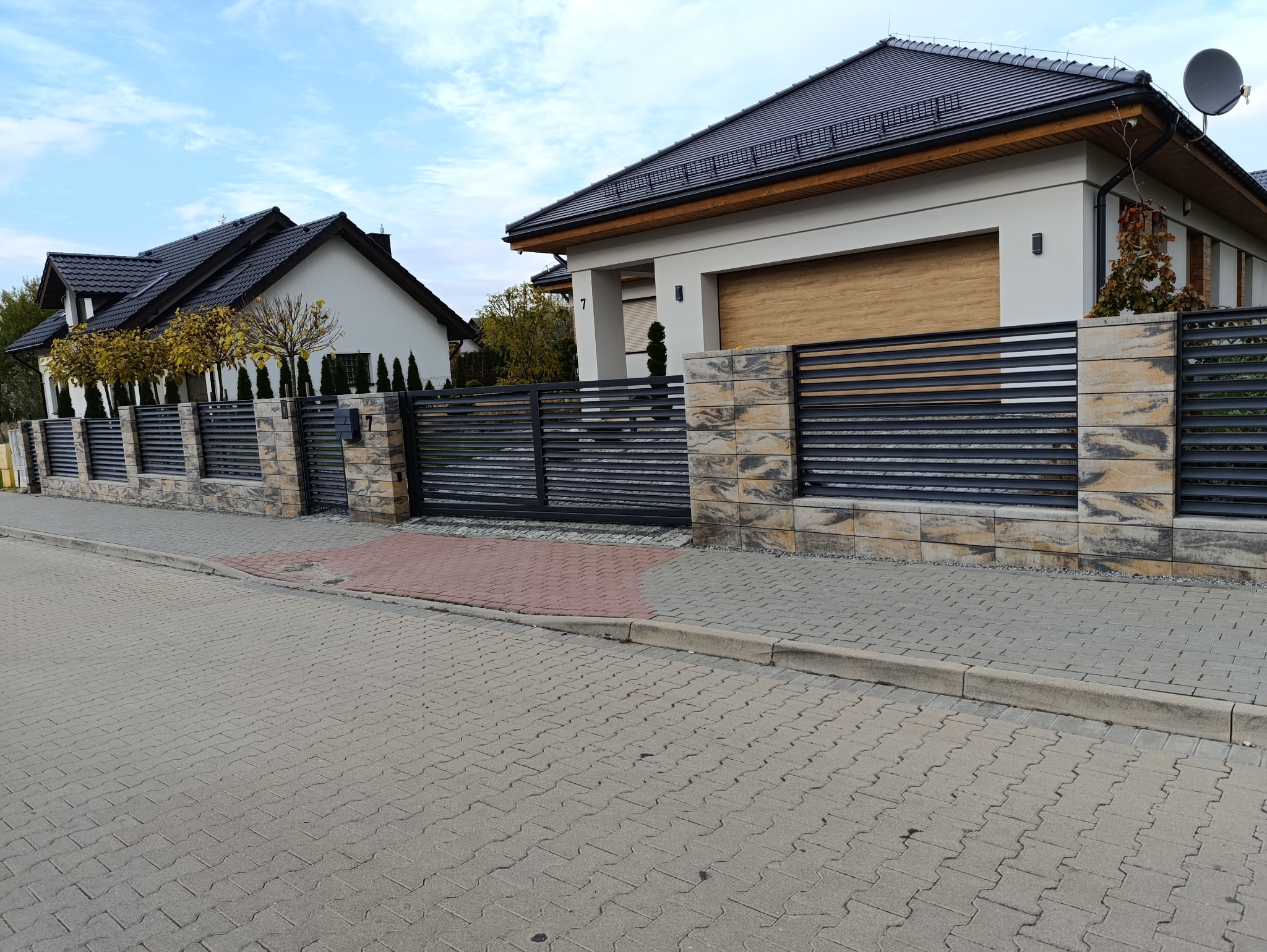 Nowoczesne ogrodzenie panelowe z elementami imitującymi kamień, oddzielające posesję od ulicy. Widoczne dwie bramy wjazdowe i furtka, dom w tle.