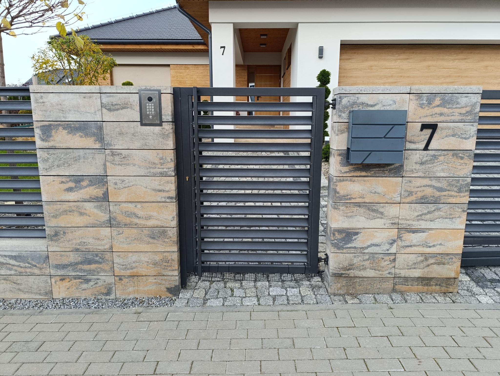 Nowoczesna brama z poziomych profili, osadzona między słupkami z bloczków w odcieniach beżu i szarości. Widoczny dom z numerem 7, skrzynka na listy i domofon.