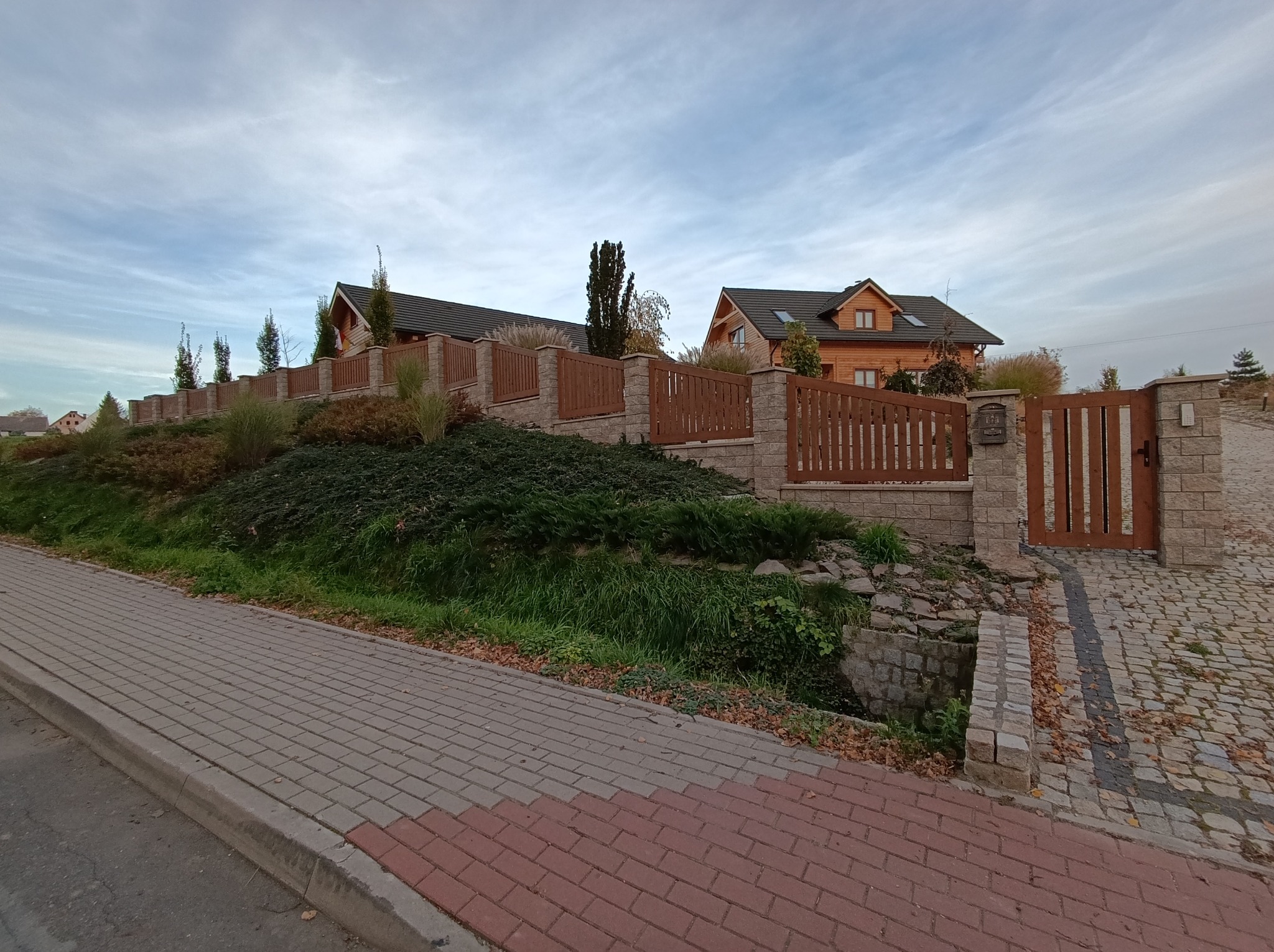 Drewniano-kamienne ogrodzenie posesji z bramą wjazdową, na tle drewnianych domów. Kątowe ujęcie z poziomu chodnika z kostki brukowej.
