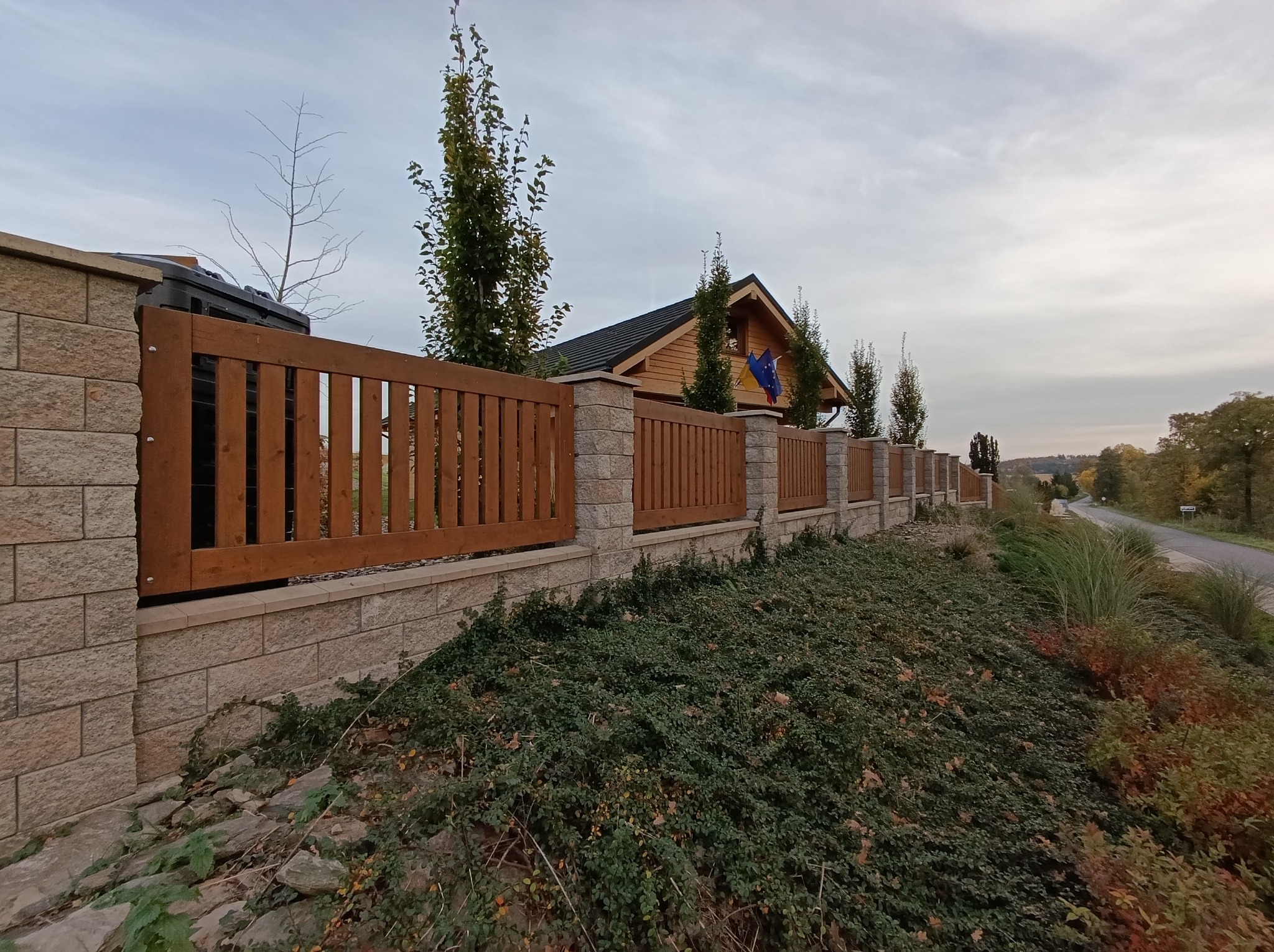 Długie, drewniano-kamienne ogrodzenie posesji z widocznym budynkiem z flagami, zieleń na pierwszym planie, pochmurne niebo.