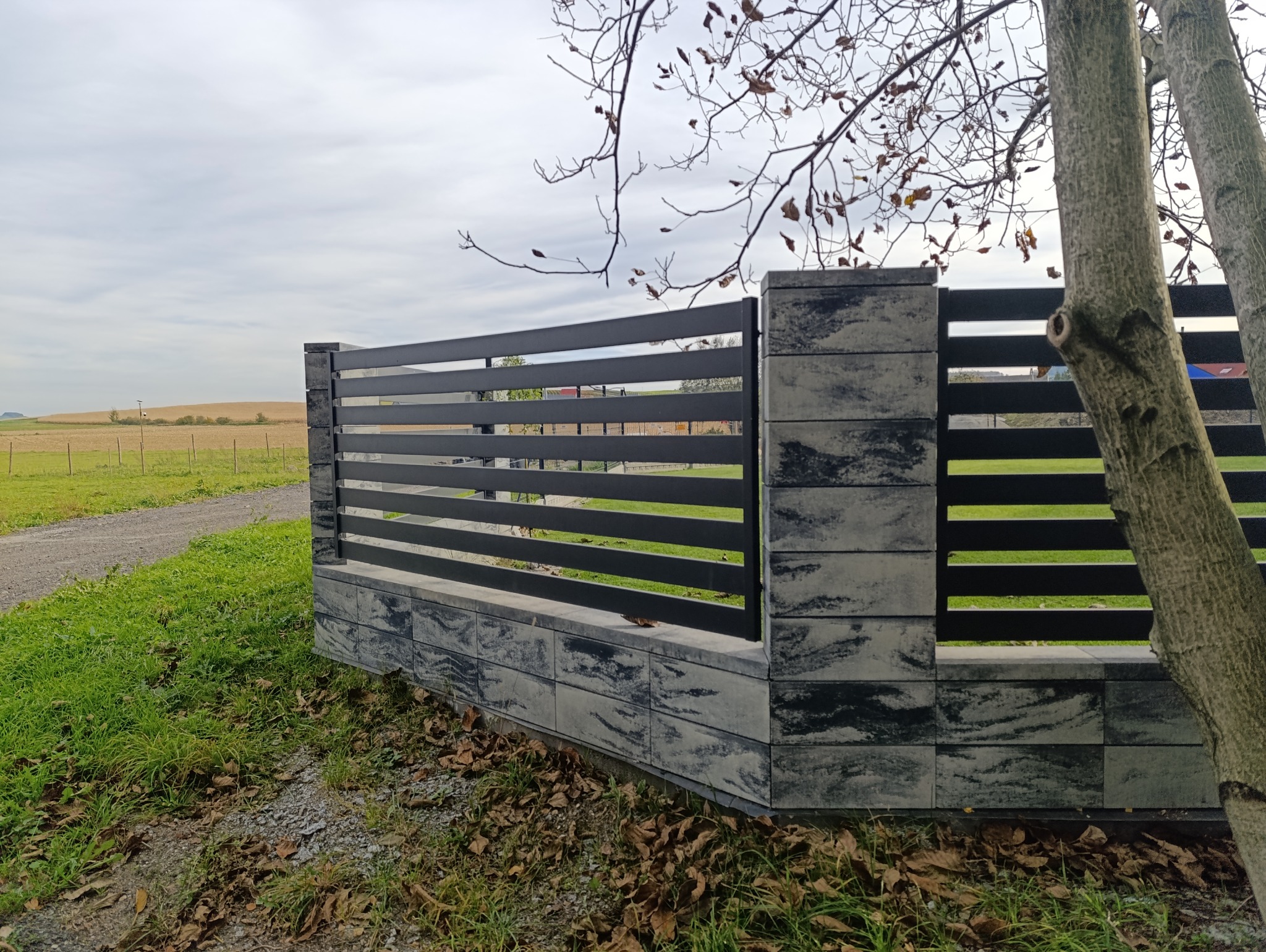 Nowoczesne ogrodzenie betonowe z poziomymi, czarnymi przęsłami, widok z boku. Tło: zielone pole i drzewa. Fragment pnia drzewa w prawym dolnym rogu kadru.