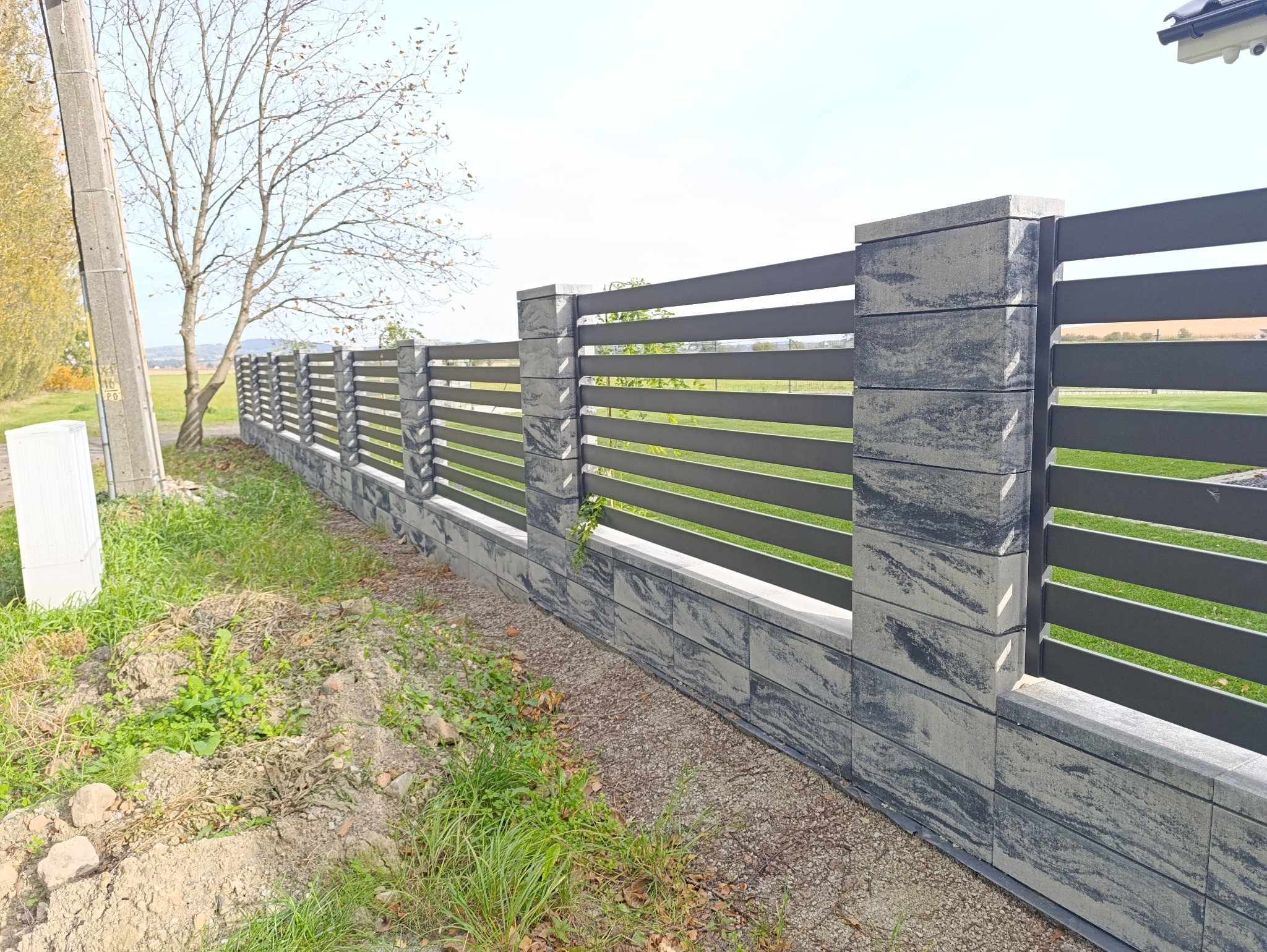 Nowoczesne ogrodzenie z szarego betonu architektonicznego z poziomymi, metalowymi przęsłami. Widok na fragment posesji i drzewo w tle.